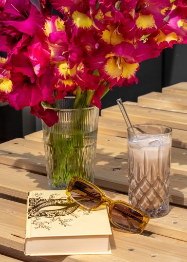Pinke Gladiolen von Beckmanns Blumen blühen in einer Vase. Daneben stehen ein Eiskaffee, ein Buch und eine Sonnenbrille.