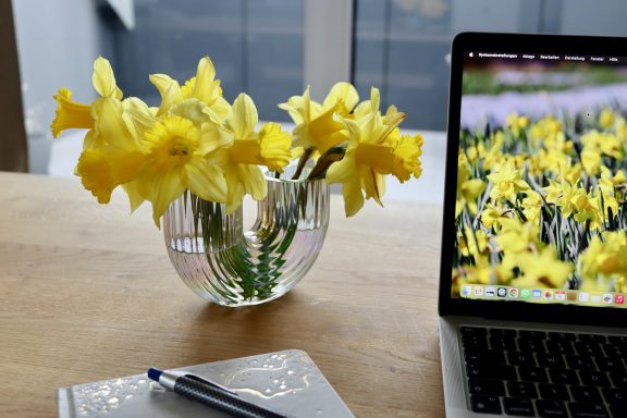 Gelbe Narzissen von Beckmanns Blumen blühen in einer Vase neben einem Laptop und einem Notizbuch auf einem Schreibtisch.
