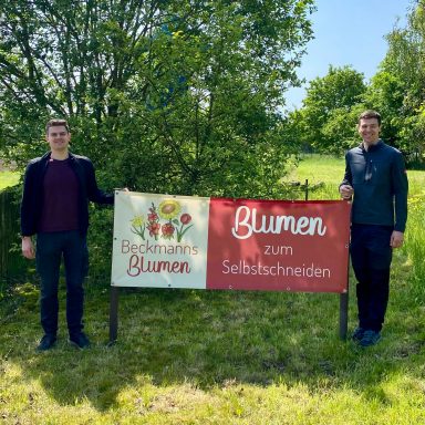 Wilhelm und Justus Beckmann von Beckmanns Blumen stehen neben einem Schild für "Blumen zum Selbstschneiden".